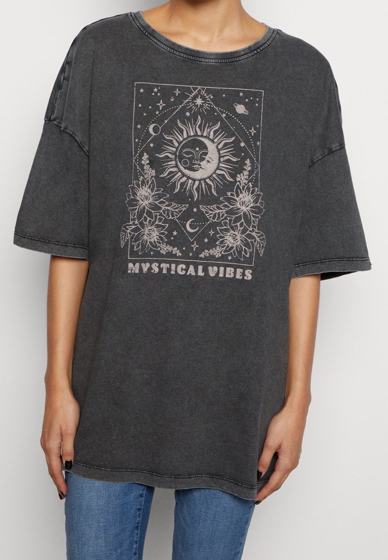 Personne portant un t-shirt noir délavé, oversized, avec un motif de soleil, lune, étoiles, fleurs, et le texte « Mystical Vibes » en impression claire sur le devant.