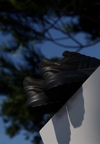 Pair of black athletic shoes resting on a white surface with blurred trees and blue sky in the background.
