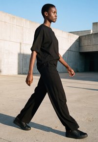 Henkilö kävelee ulkona betoniseinien vieressä päällään väljä musta t-paita, leveät mustat housut ja mustat lenkkarit, kirkkaan taivaan alla.