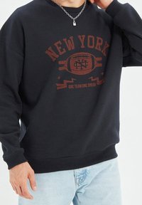 Sudadera negra con la inscripción "NEW YORK" en letras rojas, gráfico centrado de un balón de fútbol, puños y dobladillo acanalados, combinada con jeans de color azul claro.