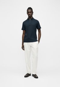 Jeune homme debout, les mains dans les poches, portant une chemise à col zippé manches courtes bleu marine foncé, un pantalon blanc et des chaussures marron.