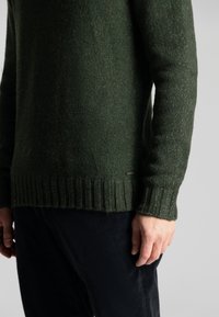 Dunkelgrüner Strickpullover mit gerippten Bündchen und Saum, der eine strukturierte Oberfläche aufweist. Der Pullover wird mit dunklen Cordhosen kombiniert.