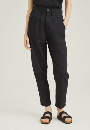 Personne portant un pantalon noir taille haute fuselé avec des plis, un haut noir, des sandales noires à double bride et une bague à la main gauche.