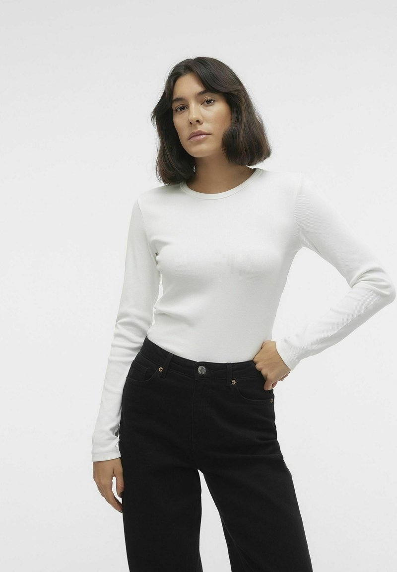 Witte langslavende geribde top gecombineerd met hoge zwarte jeans. De broek heeft subtiele zakken en een metalen knoopdetail.
