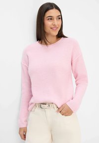Hellrosa gestrickter Pullover mit Rundhalsausschnitt, überschnittenen Schultern und gerippten Bündchen, kombiniert mit cremefarbener Hose und einem Gürtel.