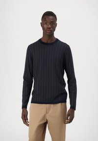 Maglione a maglia blu navy con motivo a spina di pesce, scollatura rotonda e maniche lunghe. Indossato con pantaloni beige, presenta una finitura testurizzata.