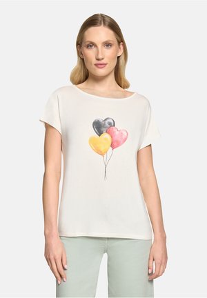 Vrouw draagt een wit shirt met korte mouwen met zwarte, roze en gele hartvormige ballonnen op de voorkant, gecombineerd met lichtgroene broek.