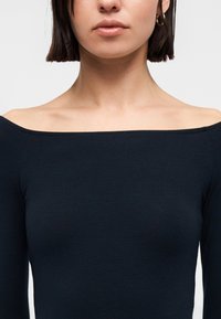 Γυναίκα με ίσια καστανά μαλλιά, που φορά χρυσούς κρίκους στα αυτιά και μαύρη μπλούζα με μακριά μανίκια που αφήνει τους ώμους ακάλυπτους, σε λευκό φόντο.