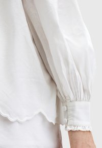Blusa bianca con maniche voluminose, orlo scallop e polsini a volant. Il tessuto appare leggero e leggermente testurizzato. Accentuata con bottoni.