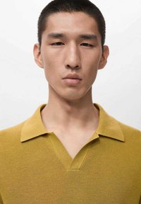 Polo giallo senape con un tessuto lavorato a maglia, caratterizzata da una scollatura a V e un colletto strutturato. Design minimalista su uno sfondo semplice.