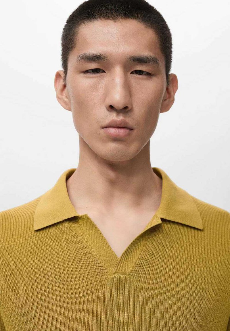 Senfgelbes Polohemd mit strukturiertem Strickmuster, ausgestattet mit V-Ausschnitt und strukturiertem Kragen. Minimalistisches Design vor einfarbigem Hintergrund.