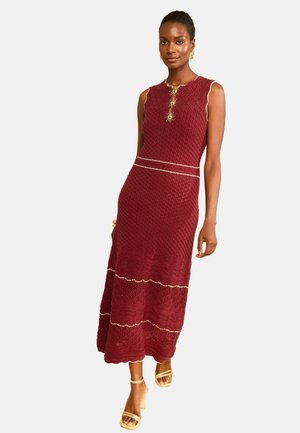 Moteris vilki bordo spalvos be rankovių megztą ilgą suknelę su tekstūriniais raštais, dekoratyviomis auksinėmis detalėmis, auksiniais auskarais, apyranke ir auksiniais basutėmis su dirželiais.