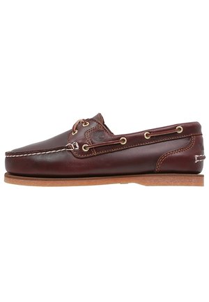 Ciemnobrązowy skórzany but typu boatshoe z beżowymi przeszyciami, sznurowadłami przechodzącymi przez metalowe oczka i płaską jasnobrązową podeszwą, widoczny z boku.