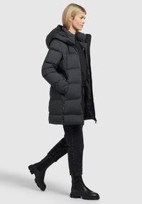 Doudoune noire avec capuche, présentant un design matelassé et une fermeture éclair à l'avant. Assortie à un jean noir et des bottines à hauteur de cheville.