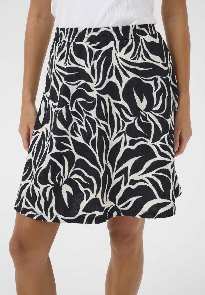 Femme portant une jupe noire et blanche à motifs floraux longueur genou avec une ceinture élastique et un haut blanc.