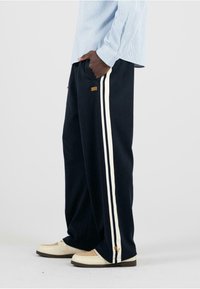Die marineblauen Jogginghosen haben weiße Seitenstreifen und ein kleines goldenes Emblem. Kombiniert mit cremefarbenen Sneakern und einem hellblauen gestreiften Hemd.