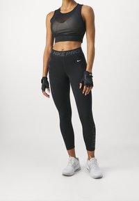 Nainen, joka on pukeutunut mustiin Nike Pro -leggings-housuihin, mustaan lyhyeen urheilupaitaan, sormettomiin hanskoihin ja valkoisiin urheilukengiin, seisoo valkoista taustaa vasten.