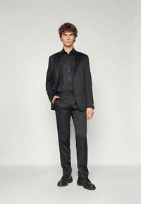 Jaunas vyras, vilkintis tamsiomis raštuotomis kostiumo kelnėmis ir juoda marškiniais bei juodais batais, stovi priešais vientisą šviesų foną.