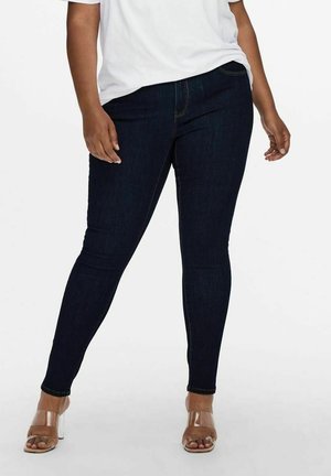 Osoba ubrana w ciemnoniebieskie dopasowane dżinsy, biały top i sandały na szpilce z przezroczystymi paskami, stojąca z jedną nogą lekko wysuniętą do przodu.