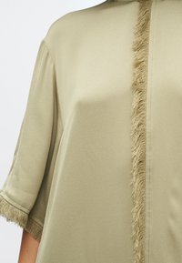 Svetlo olivovo zelená blúzka s krátkym rukávom, ktorá má jemný fringing detail na prednej strane. Hladká textúra s voľným strihom.