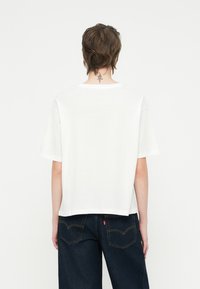 T-shirt branca de algodão com um corte descontraído, mangas largas e decote redondo. A parte de trás apresenta detalhes minimalistas e um acabamento de bainha subtil.