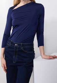 Vrouw in een strakke marineblauwe lange mouw top en donkerblauwe high-waisted jeans, leunend op een witte ondergrond met één hand.