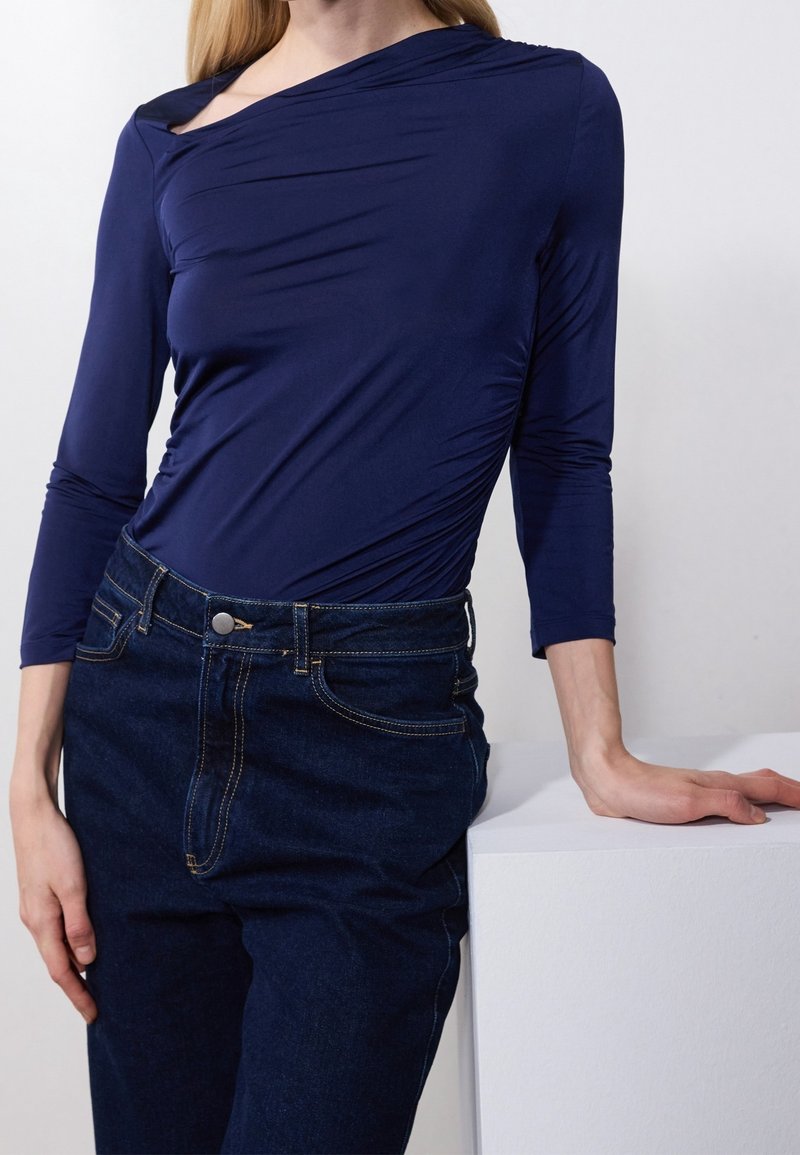 Vrouw in een strakke marineblauwe lange mouw top en donkerblauwe high-waisted jeans, leunend op een witte ondergrond met één hand.