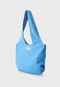 Μπλε τσαντάκι ώμου από καμβά με φαρδιά τιράντα, σε ορθογώνιο σχήμα και μικρή ετικέτα με λογότυπο. Διαθέτει κόκκινες λεπτομέρειες στις πλευρές.