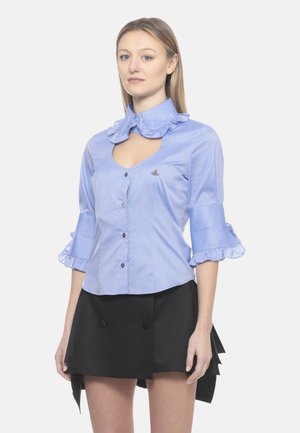 Donna che indossa una camicia celeste con colletto e maniche arricciate, abbinata a una gonna nera asimmetrica.