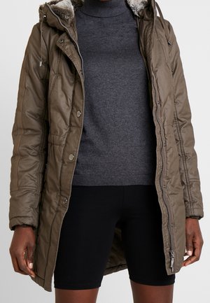 Personne portant un manteau matelassé olive longueur genoux avec une capuche doublée de fourrure, un pull col roulé gris foncé et un short noir.