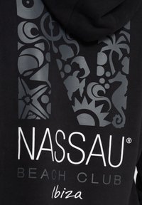 Mustat hupparit, joissa on suuri graafinen koriste, jossa on monimutkaisia harmaita kuvioita ja symboleita, mukaan lukien meritähti, merenhevosia ja trooppisia motiiveja. Lisäksi tekstit "NASSAU BEACH CLUB" ja "Ibiza" valkoisella tekstillä.