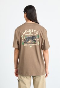 Brun bomull T-skjorte med et grafisk motiv av en svart panter, teksten "DICKIES WORKWEAR" over og "QUALITY MADE" under, med grønne blomsterakenter.