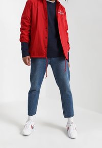 Czerwona nylonowa kurtka ze sznurkiem, nałożona na ciemnoniebieski sweter z dzianiny. Noszona z jasnoniebieskimi zwężanymi dżinsami i białymi trampkami z czerwonymi akcentami.