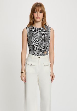 Femme portant un haut sans manches noir et blanc à imprimé zèbre avec des boutons sur le devant, et un pantalon blanc taille haute à jambes larges avec des poches à rabat.