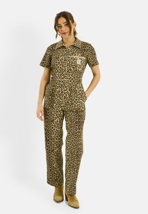 Mujer de pie con mono de manga corta con estampado de leopardo, bolsillos delanteros y botas de tacón color beige, sonriendo con una mano en el bolsillo.