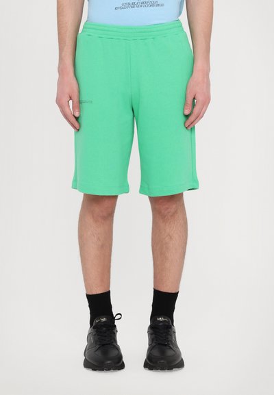 Grüne Baumwollshorts mit einem elastischen Bund, die bis zur Mitte des Oberschenkels reichen, mit Seitentaschen und einem schlichten Design. Schwarz gefärbte Sneakers getragen.