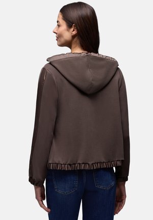 Femme aux longs cheveux bruns portant une veste à capuche marron avec des poignets et un ourlet élastiques, debout devant un fond clair uni.