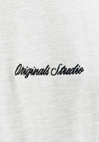 Hele halli kangas pehmea tekstuuriga, millel on musta tikandiga tekst "Originals Studio" eakate kirjas.