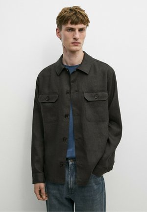 Ung mand iført en mørk jakke med knapper over en blå skjorte og blå jeans, står mod en ensfarvet hvid væg med afslappede hænder.