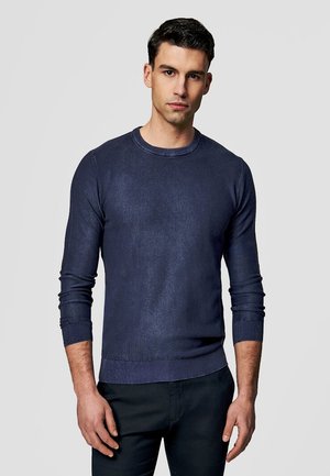 Młody mężczyzna stojący przodem, ubrany w dopasowany granatowy sweter z długim rękawem i ciemne spodnie na jednolitym jasnym tle.