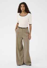 Femeie purtând un top crem cu mâneci scurte și margini negre și pantaloni bej, cu talie înaltă și picioare largi, stând cu mâinile în buzunare.