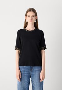T-shirt noir à manches courtes avec des finitions vertes-brunes à motifs au col et aux poignets, fabriqué dans un tissu lisse. Porté avec un jean bleu.