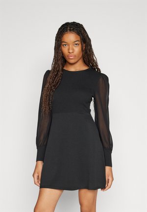 Black dress featuring a ribbed bodice, sheer long sleeves, and a flared skirt. Simple design with a classic round neckline.