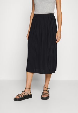 Vrouw die een zwarte geplooide midi-rok, een grijs mouwloos topje en zwarte platte sandalen met bandjes draagt, staand tegen een effen witte achtergrond.