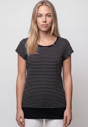 Blonde Frau mit glattem Haar, die ein schwarz-weiß gestreiftes Shirt mit kurzen Ärmeln trägt und vor einem einfarbigen Hintergrund steht.