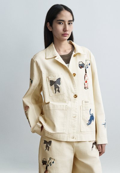 Chaqueta beige claro de algodón con diseños bordados de animales, con cuello de solapa, bolsillos delanteros y botones de madera.