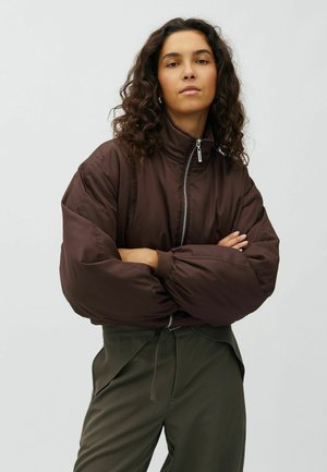 Frau mit lockigem Haar, die eine dunkelbraune Daunenjacke und olivgrüne Hose trägt, steht mit verschränkten Armen vor einem schlichten Hintergrund.