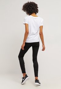 Mujer con cabello rizado, vistiendo una camiseta blanca, pantalones ajustados negros y zapatillas Nike negras, de pie frente a un fondo claro.