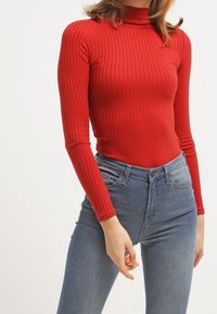 Top lungo a maniche corte rosso a coste con collo alto. Abbinato a jeans in denim blu aderenti, evidenziando una silhouette snella e un tessuto strutturato.