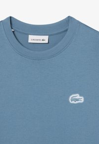 T-shirt di cotone azzurro chiaro con collo a giro, maniche corte e logo di un coccodrillo ricamato in bianco sul lato sinistro del petto.